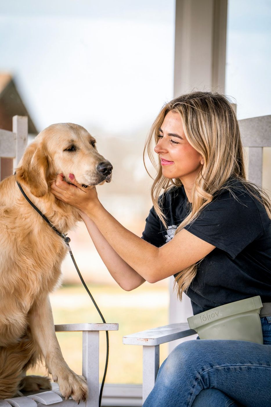 A trainer pets the face of a golden retriever on a sunny porch