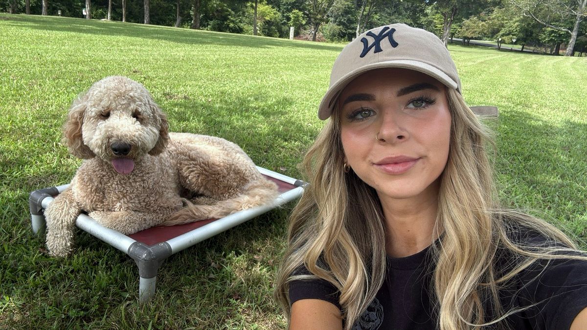 A trainer takes a selfie beside a curly goldendoodle lying on a raised dog bed on the lawn
